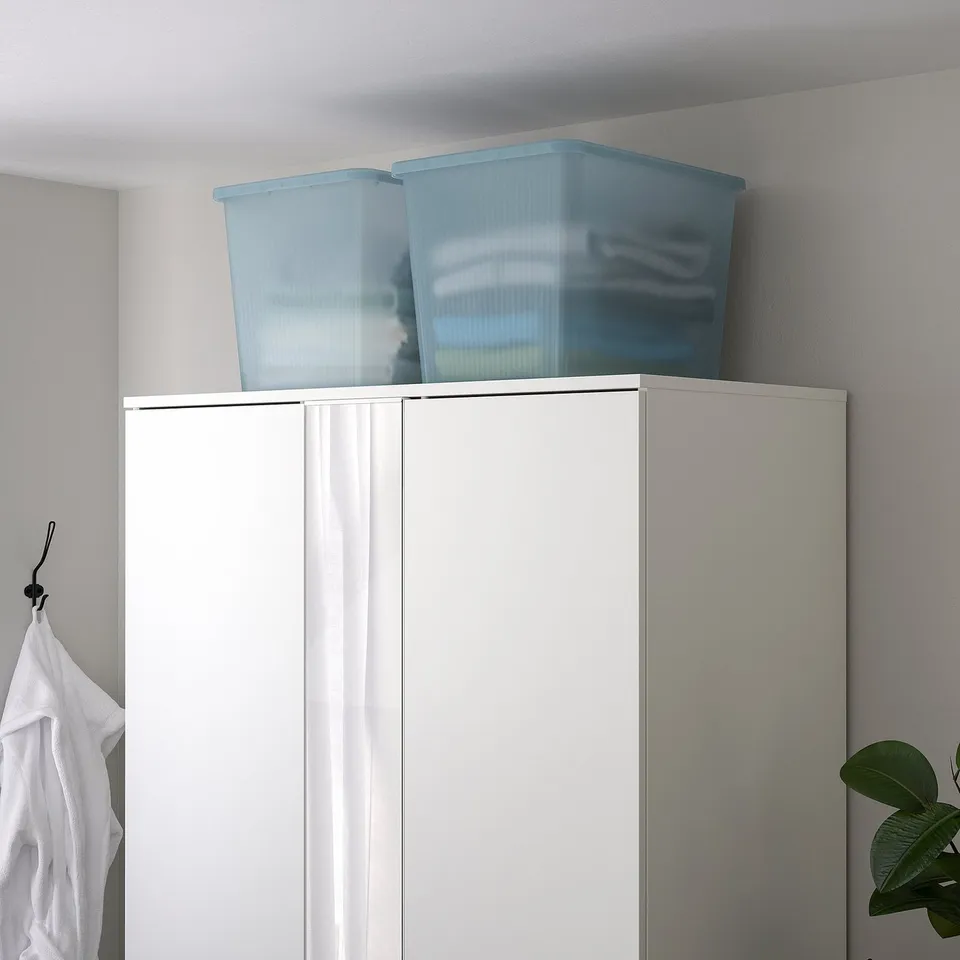 Two light blue, transparent plastic RYKTA boxes atop white wardrobe, larger than nearby white shirt.