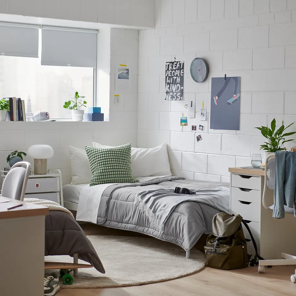 A bedroom with a LÅSBRÄKEN grey comforter on a bed, green pillow, white walls, a large window, desk with chair, plants, and posters.