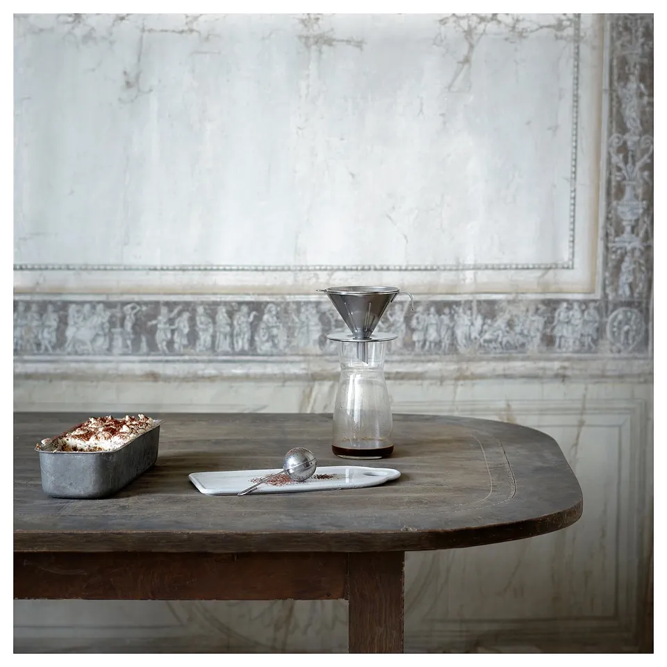 Wooden table displays a reusable metallic coffee philtre, clear glass container, and tray with coffee grounds. Elegant, minimalist setup.