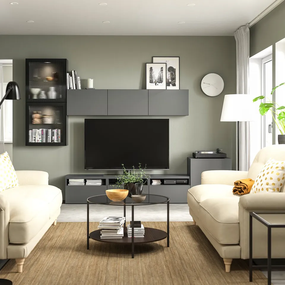 Modern living room with sage green walls, white sofas, BESTÅ tv bench, books, and a glass coffee table with a plant.