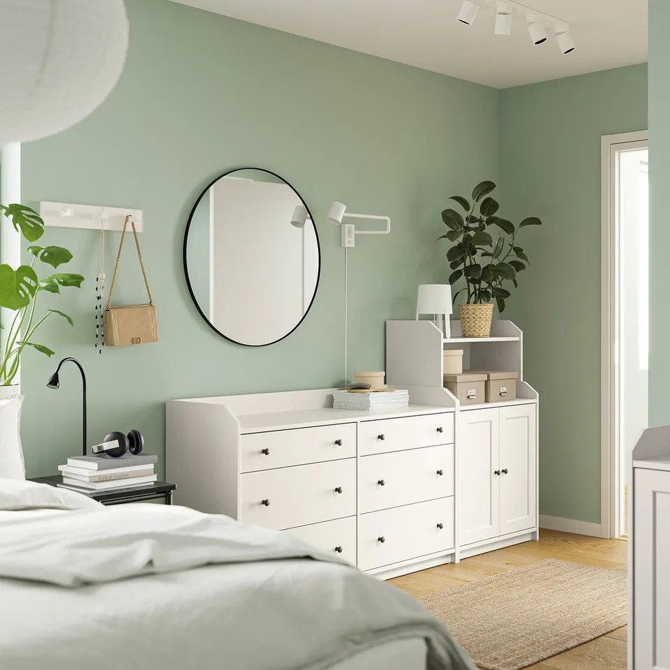 Bedroom with green walls, white HAUGA storage units, round mirror, plant, and lamp.