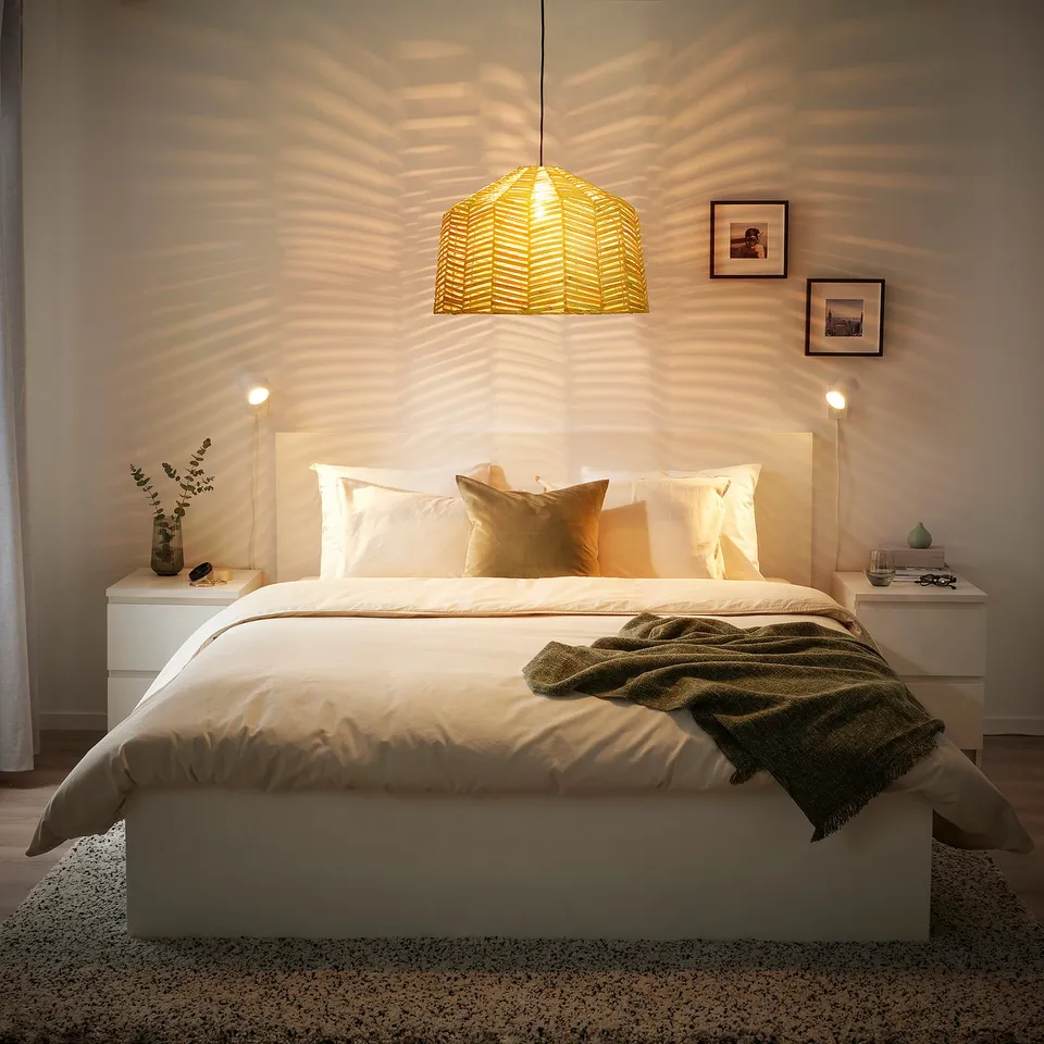 Yellow rattan KAPPELAND lamp shade casts warm light over a large, cosy bed with green blanket and pillows, contrasting with smaller bedside tables.