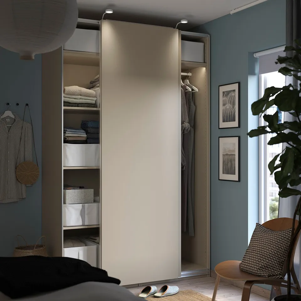 Modern bedroom with open beige PAX wardrobe, blue walls, neatly folded clothes, hanging garments, and decorative plants.