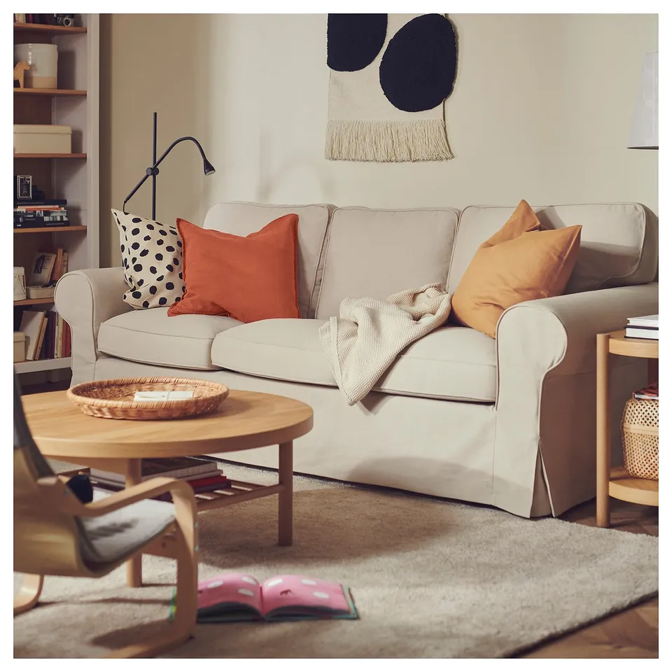Cosy living room with beige sofa, orange pillows, wooden coffee table, and fluffy rug. Wall art and bookshelf in the background.