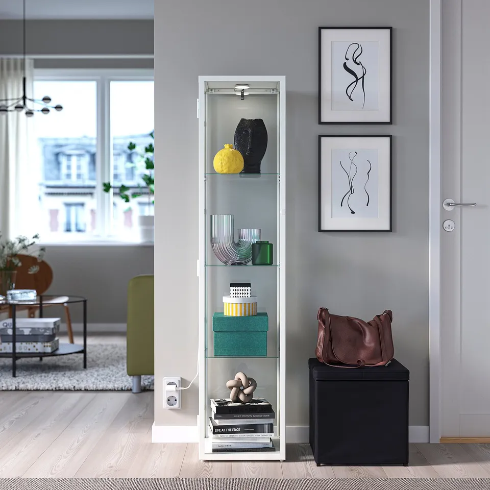 Modern living room with STRIMSÄV glass cabinet, spotlight, yellow vase, books, black side table, framed artworks, large window view of building.