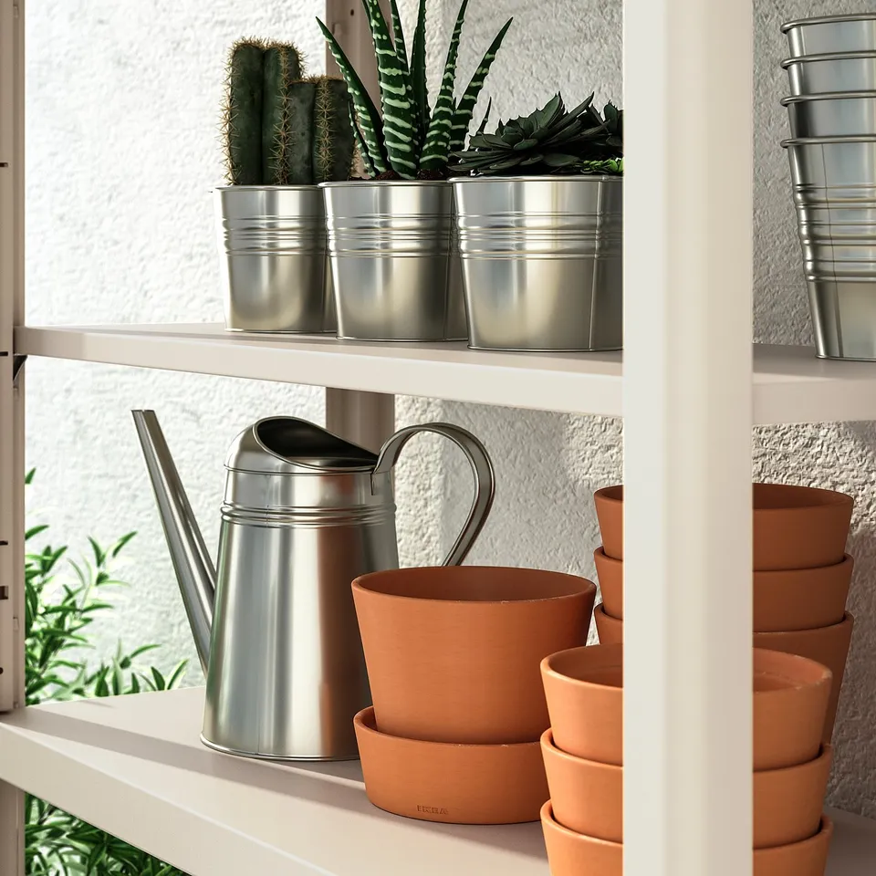 White shelf with metal watering can and terracotta pots. Silver planters on top hold cacti.
