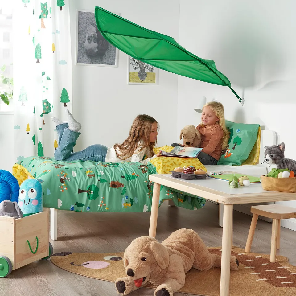 Two kids playing on a bed with a green leaf canopy; toys and blankets around.