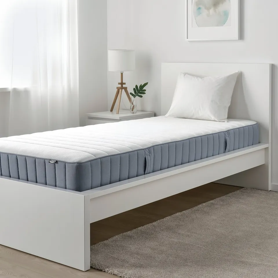 Modern bedroom with VALEVÅG firm grey mattress on white bed, single size, near small bedside table with lamp and plant, light decor.