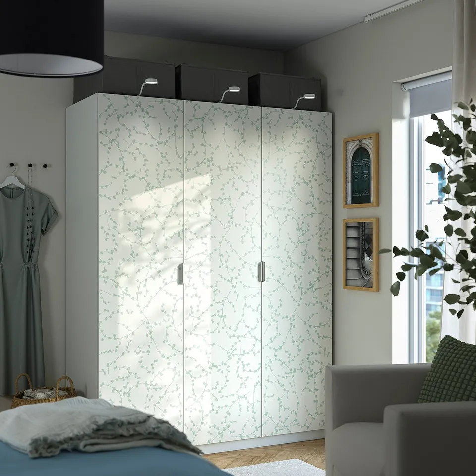 A LINNABERG white wardrobe with green leaf design, matching pax frames, placed next to a bed and chair, with a window and decorations nearby.