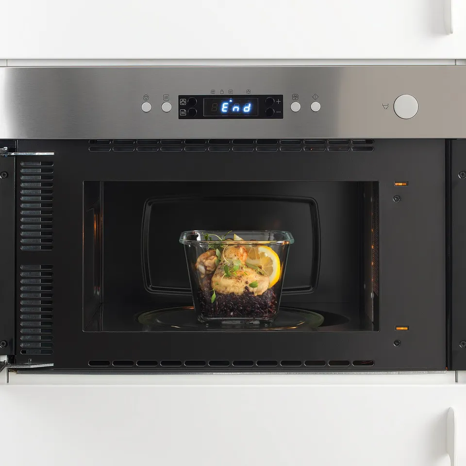 A stainless steel microwave with a digital display and a glass container inside holding cooked food.
