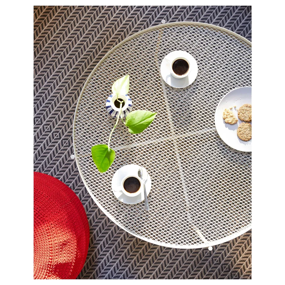 Circular outdoor table in beige has a mesh design. It holds two cups, cookies, and a small plant. A red cushion is near it.