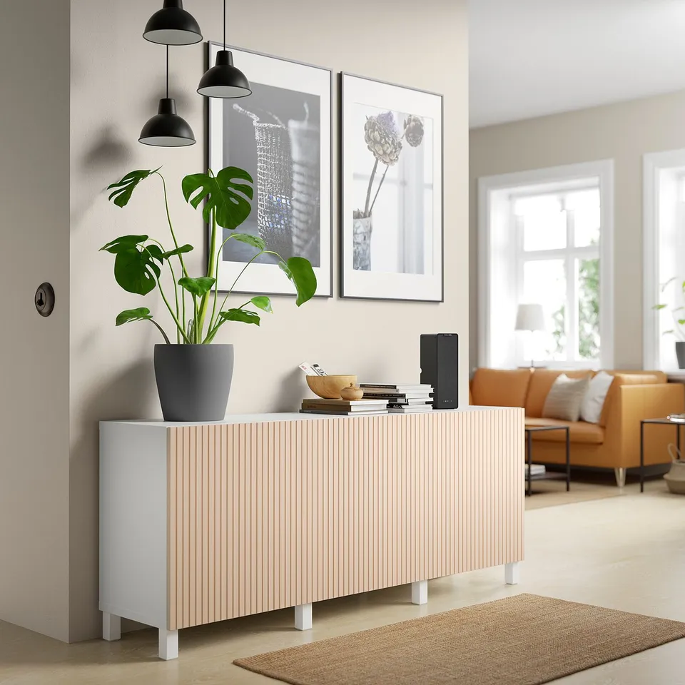 BJÖRKÖVIKEN sideboard in light birch with 3 doors, holding plant, books, and speaker. Leather furniture in background.