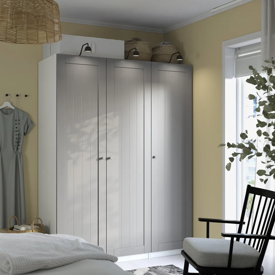 Modern bedroom with large grey PAX wardrobe. Light-colored walls, chair, and bed. Wardrobe tall, reaching ceiling. Natural light from window.