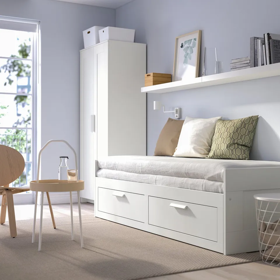 Minimalist BRIMNES day-bed with storage, beige chairs, tall cupboard, and pillows. Light blue wall, floating shelf, large window, scandinavian room.