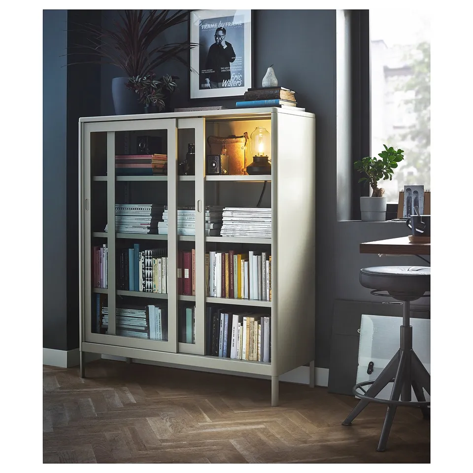 Modern home office with IDÅSEN light grey metal cabinet displaying books. Sliding glass doors, sleek design, desk with chair, and green plants.