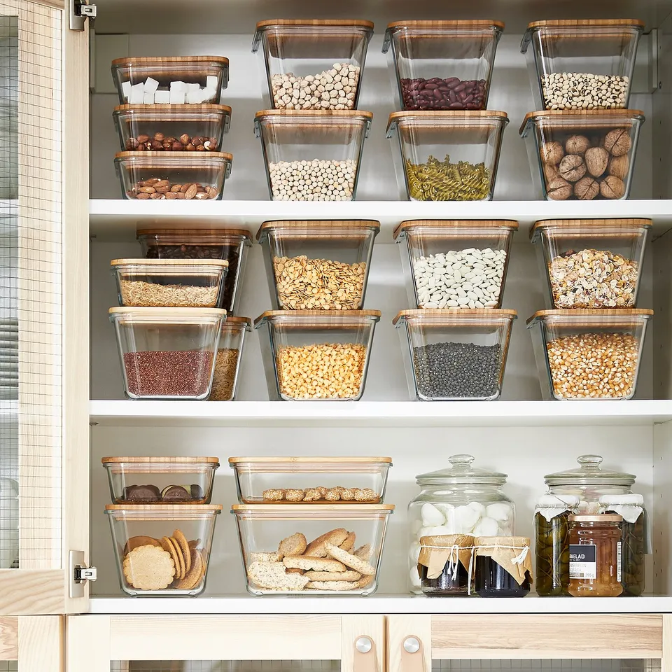 Organised kitchen shelf with clear jars storing cereals, nuts, beans, cookies. Jars have bamboo lids, grouped neatly for easy access.