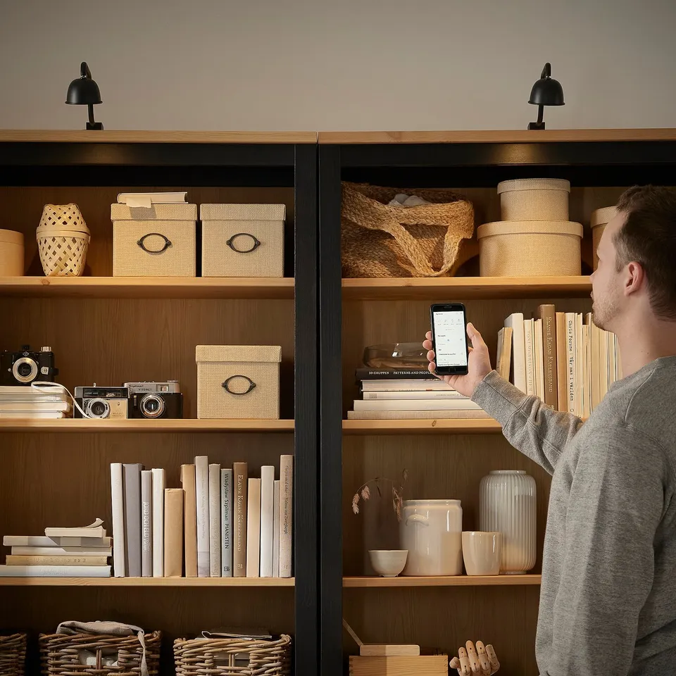 Person adjusting phone, standing before wooden cabinets with various items. Cabinets lit by two adjustable, focused lamps, highlighting contents.