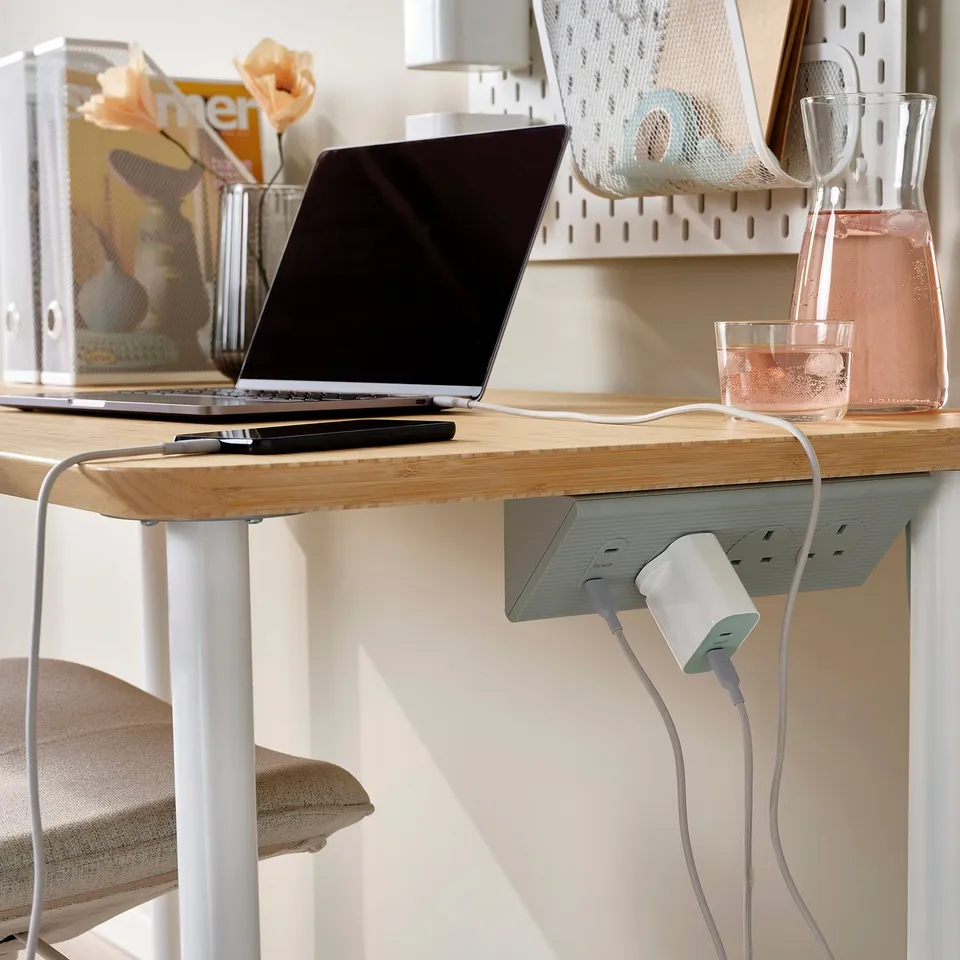 A wooden desk with a laptop, notepad, and charging station featuring multiple sockets and usb ports, with a trailing white cable.