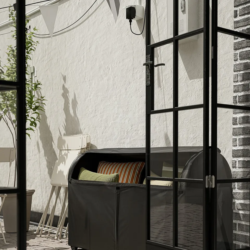 Black waterproof storage box with cushions inside, placed near a white wall with glass doors. Its designed for outdoor use and easy to move.