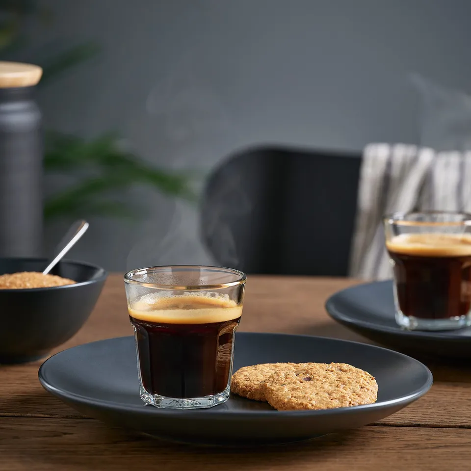 Two espresso glasses with froth, one with cookie, steam rising, sugar bowl, plant in background.