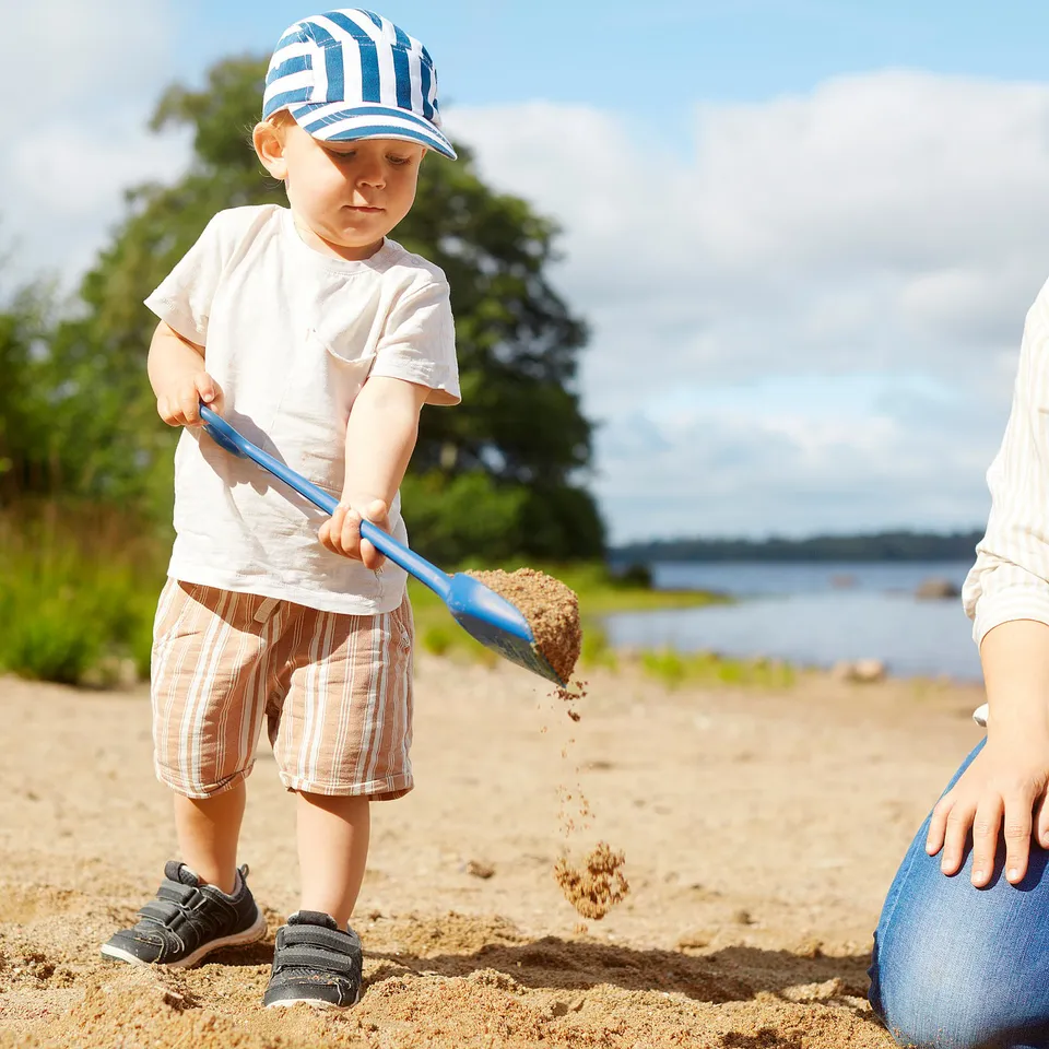 Child plays with blue spade on sandy beach, digging sand, an adult nearby.