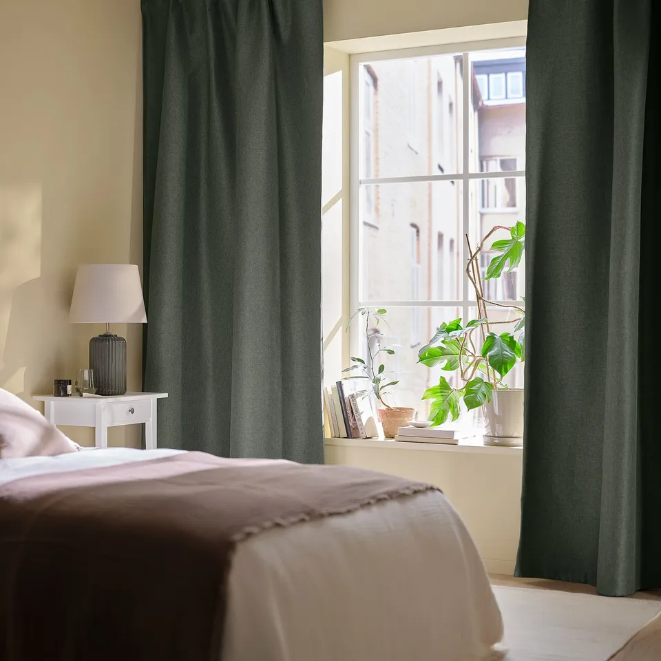 Green curtains in bedroom, drawn to side, framing window with building view. Bed with beige bedding, plants, books, and lamp on bedside table.