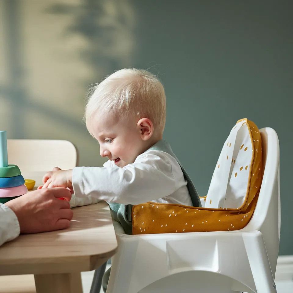 A baby sits securely in a white high chair with an inflatable yellow and white dotted cushion, learning to eat safely at a wooden table.