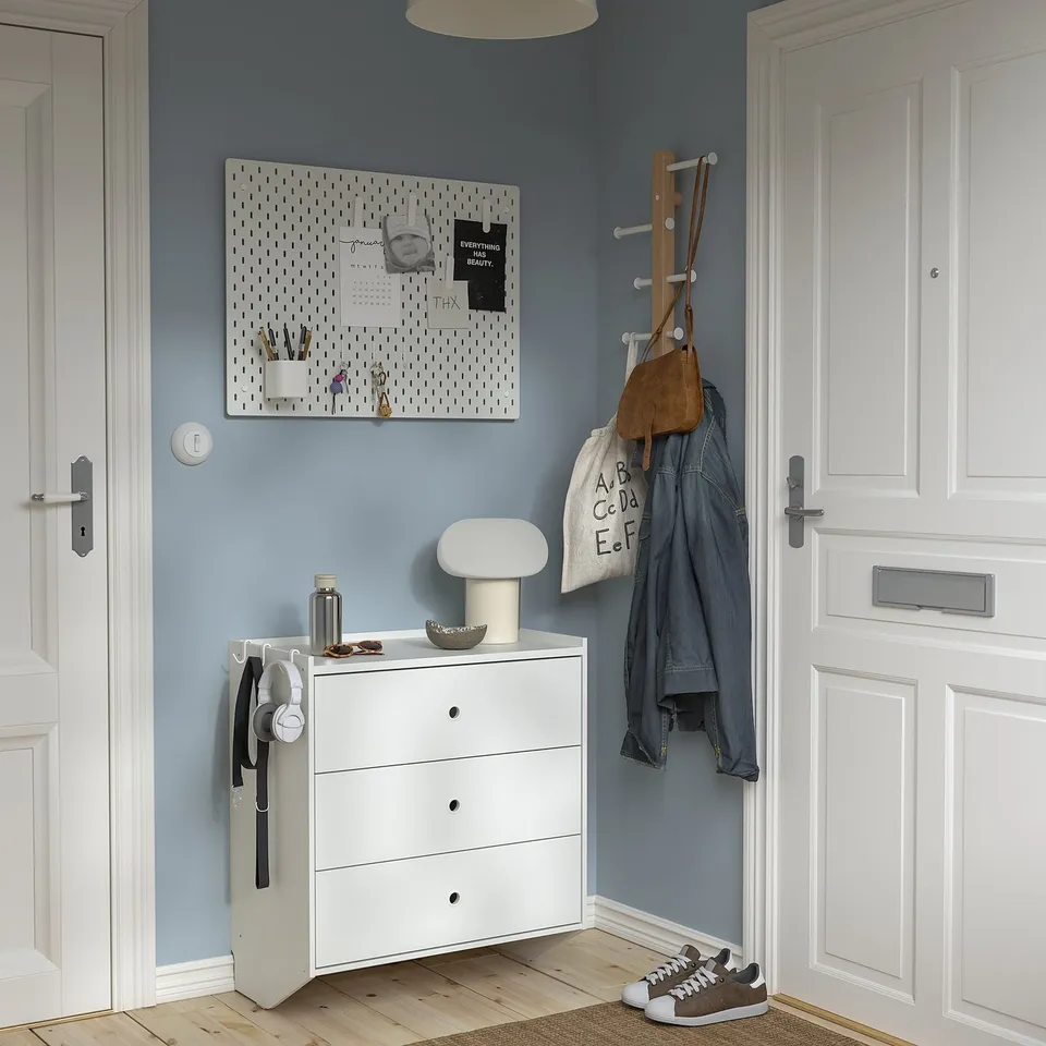 White RACKNÄS chest with 3 drawers, coat rack above, blue wall, pegboard, lamp, bowl, keys.