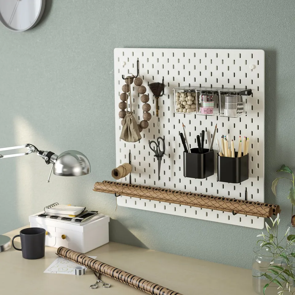 SKÅDIS pegboard organiser on green wall holds cups, brushes, scissors, containers, and a lamp above desk.