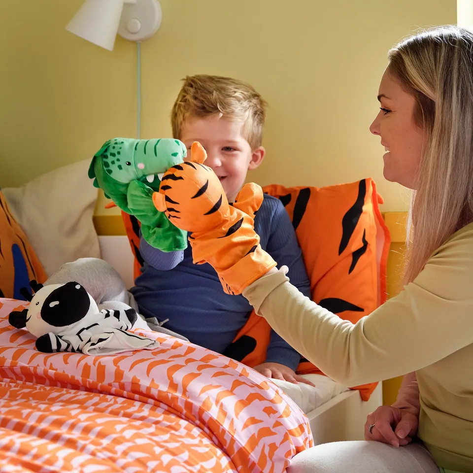 Child & adult play with tiger & crocodile puppets on a tiger-patterned NATTHÄGER bed.