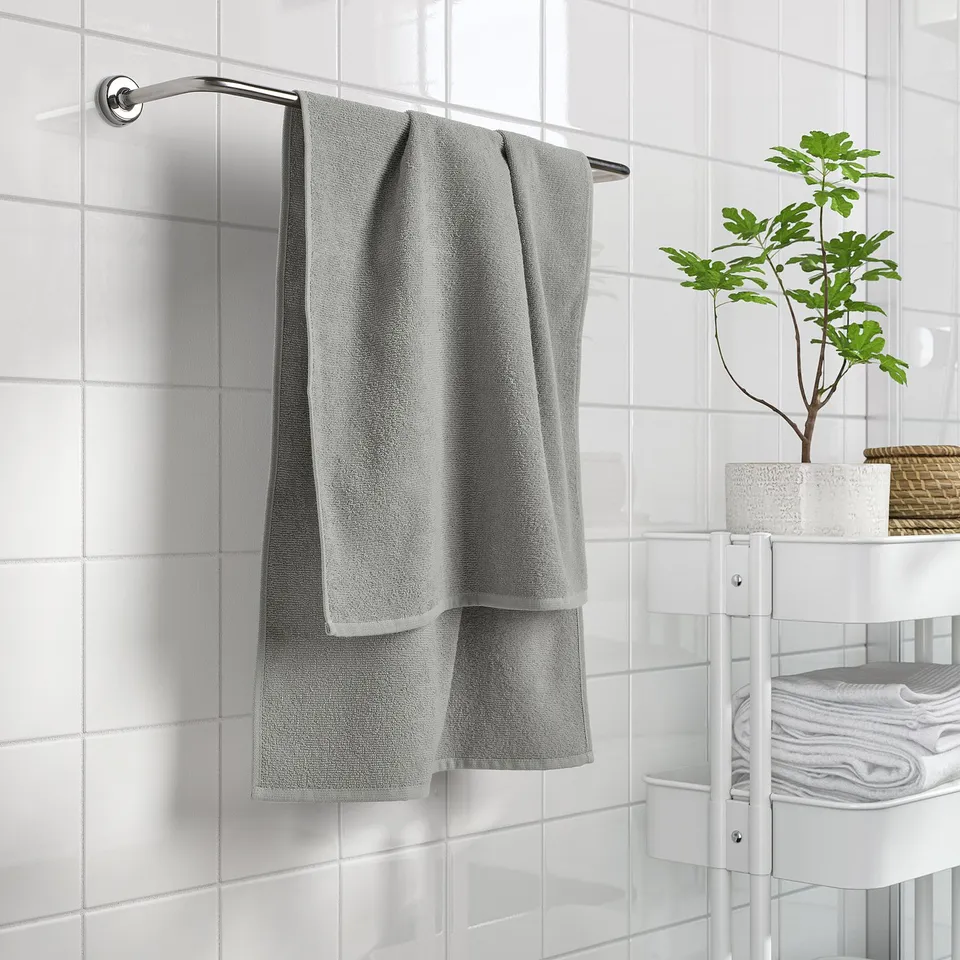 A grey LUDDVIAL towel hangs near white-tiled walls, smaller than stacked towels on a shelf with a potted plant.