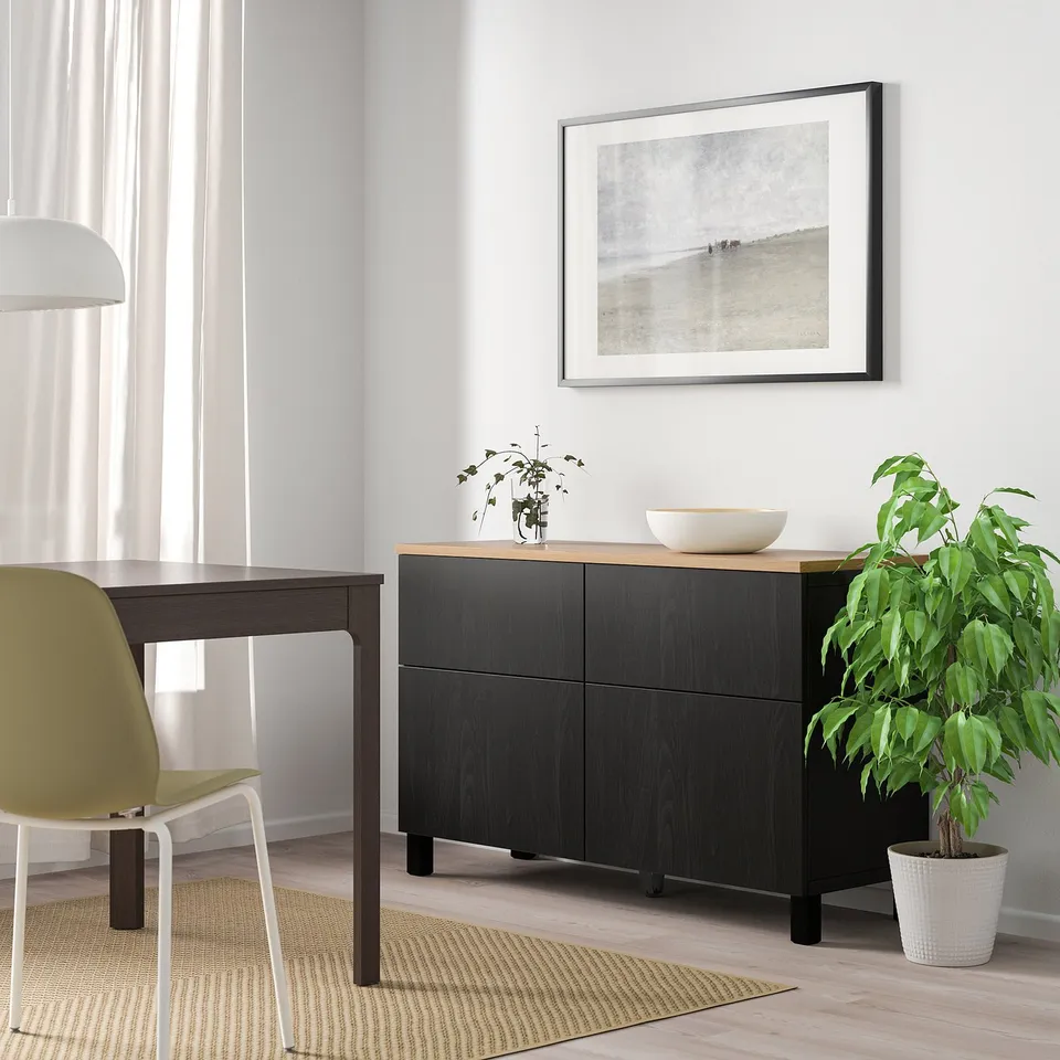 Minimalist dining setup: black sideboard, green plant, yellow chair, with framed art.
