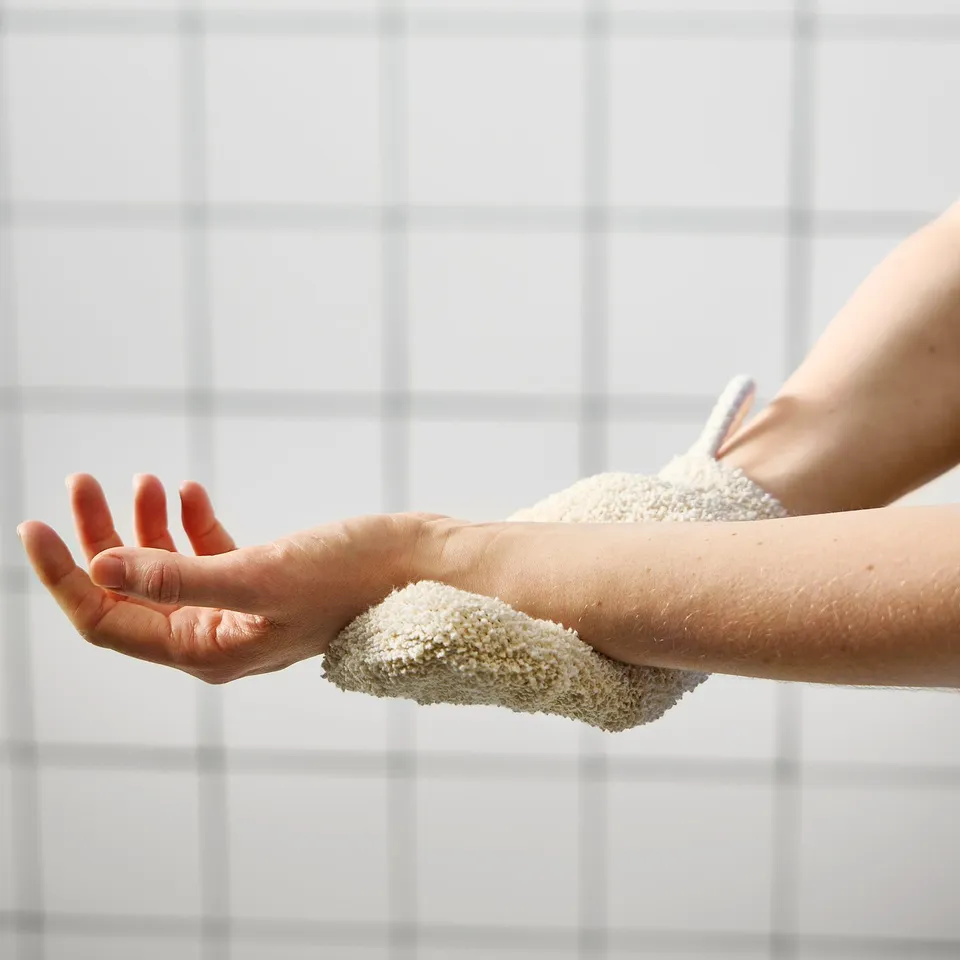 Person using a soft mesh scrub mitt with soap to clean their arm against a tiled wall.