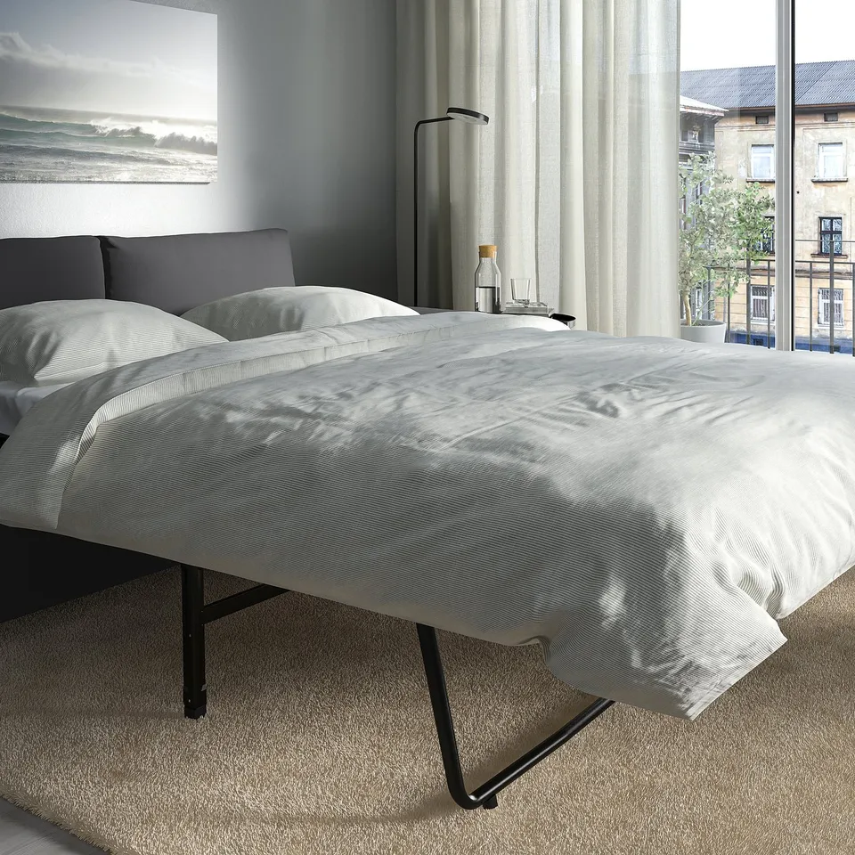 Modern bedroom with VIMLE sofa-bed, large window, soft white bedding, dark cushions, floor lamp, city view.