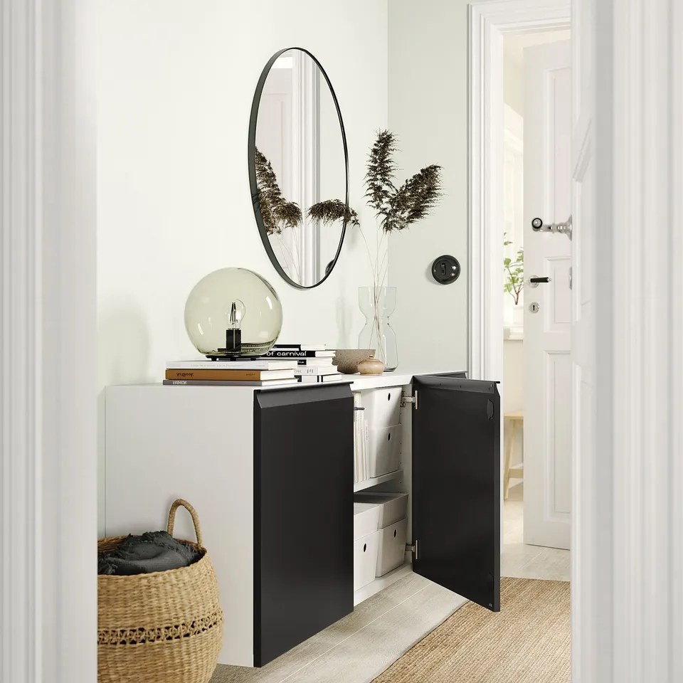 Modern entryway with white BESTÅ cabinet, black doors, round mirror, lamp, and basket.