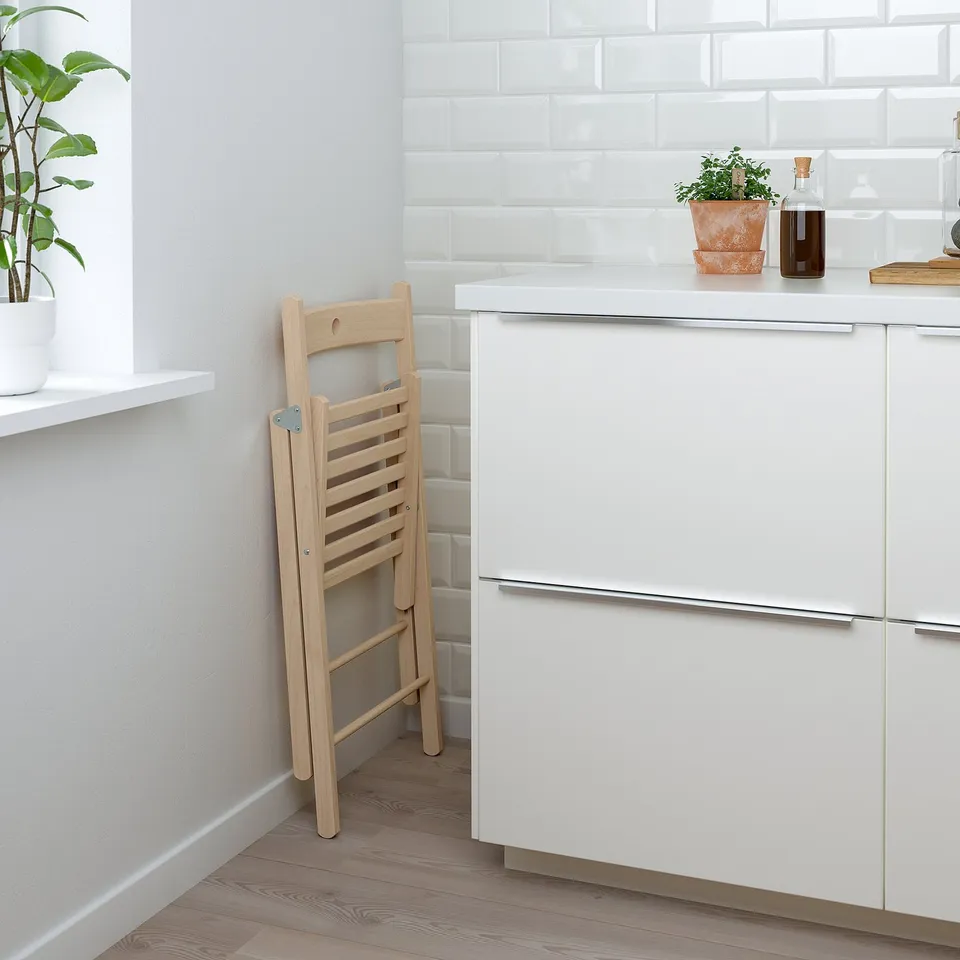 Wooden foldable kitchen chair neatly stored, leaning against wall near dishwasher in modern, vibrant kitchen with plants.