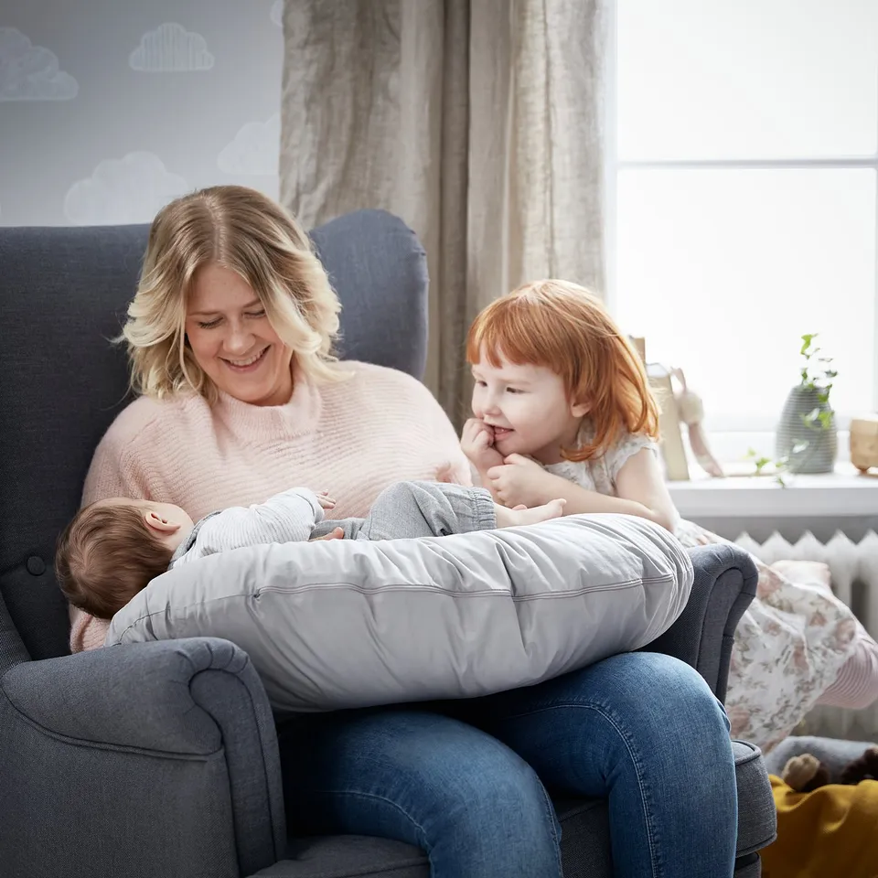 A person with two kids on a grey couch; shes smiling, holding a baby, and a red-haired girl sits beside the person.
