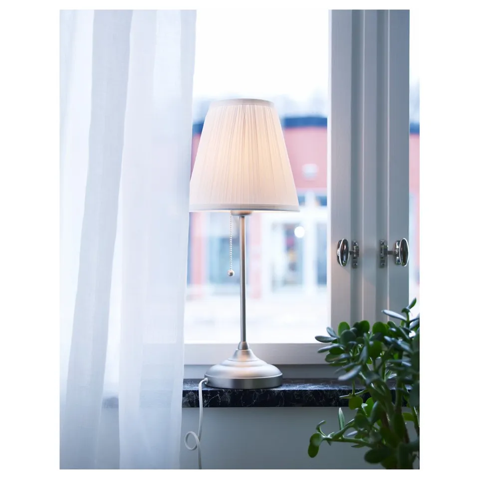 A lamp on a windowsill, with soft white shade, cord hanging down, beside a green plant.