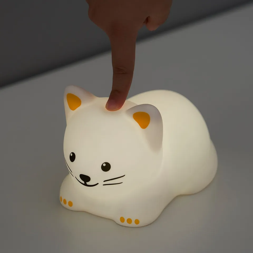 A hand touches a TÖVÄDER night light, a white cat-shaped lamp with orange accents, changing colours.