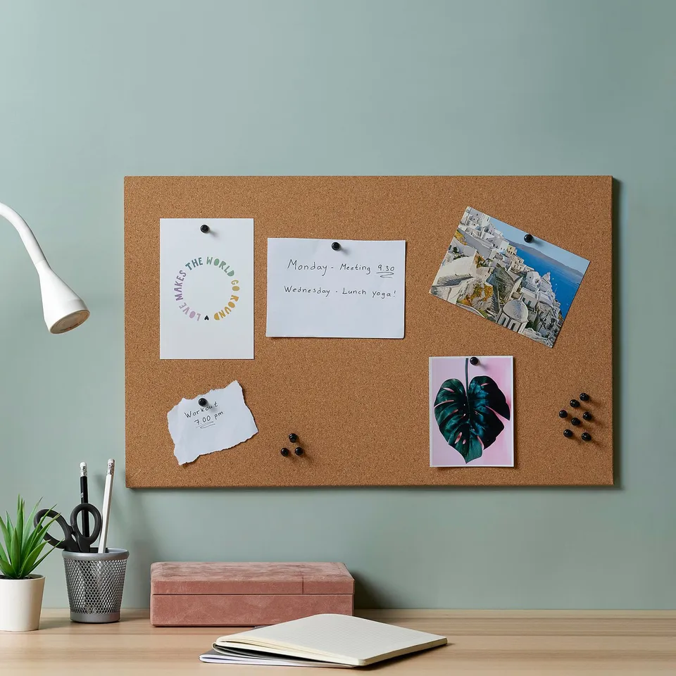 Large FLÖNSA cork memo board on wall with photos, notes, & push pins. Includes a white lamp, plant, & stationery on desk below.