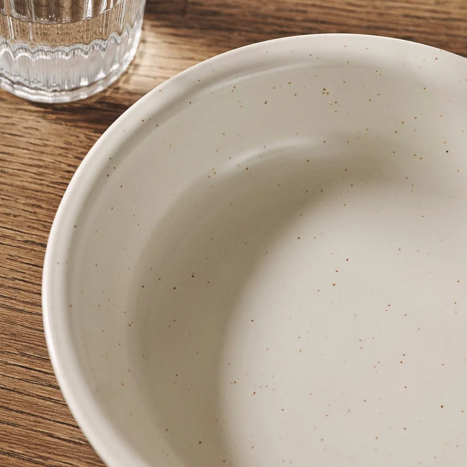A rustic beige bowl with speckled pattern sits on a wooden table, showcasing its natural, handmade texture.