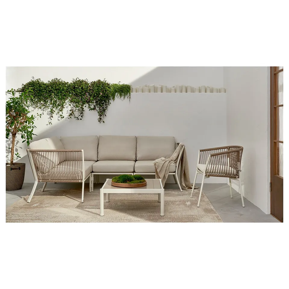 Modern patio furniture set: beige sofa, wicker chairs, glass table, white walls, green plants, neutral rug.