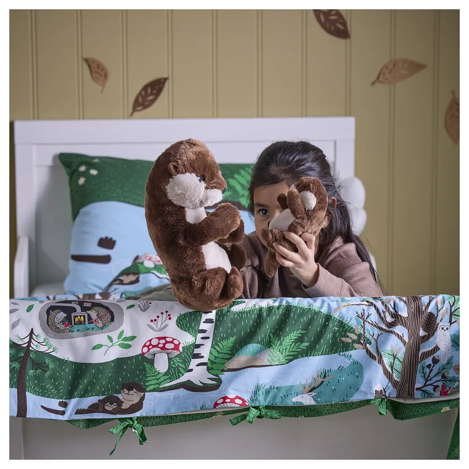 A child sits in a bed holding two plush otters, part of the SKOGSDUVA collection. The bedding a forest theme with mushrooms and otters.