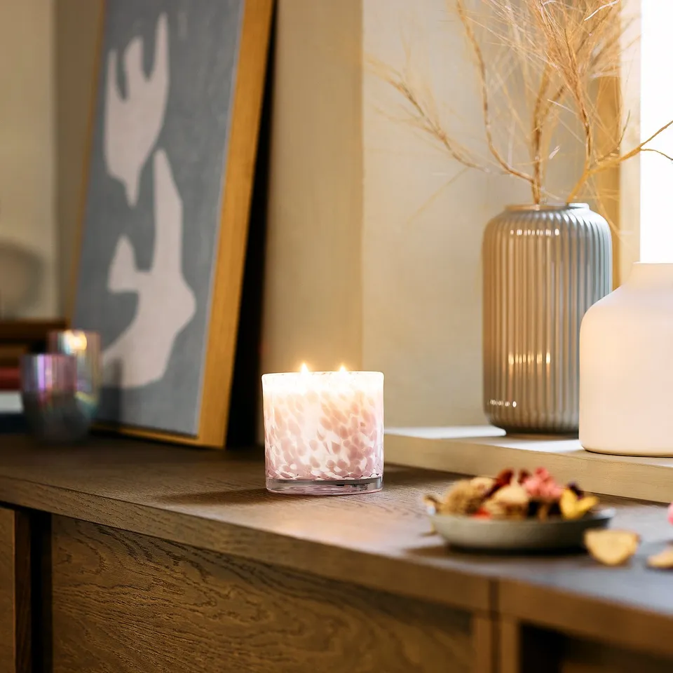 KRONSÅNGARE pink candle in glass, lit, placed on wood shelf near vase, bowl, and art. Small candle beside large items.
