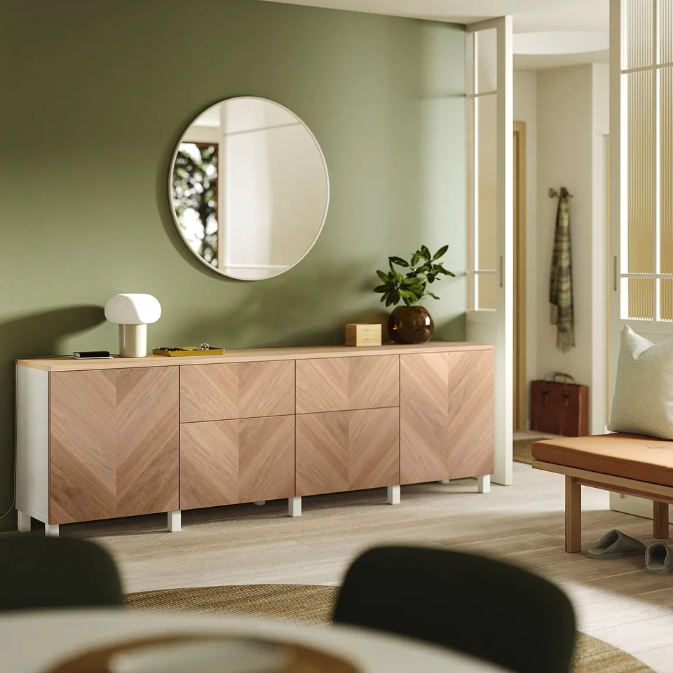 Large light wood BESTÅ cabinet against a green wall. Topped with a potted plant and small lamp. Mirror above and a bench nearby.