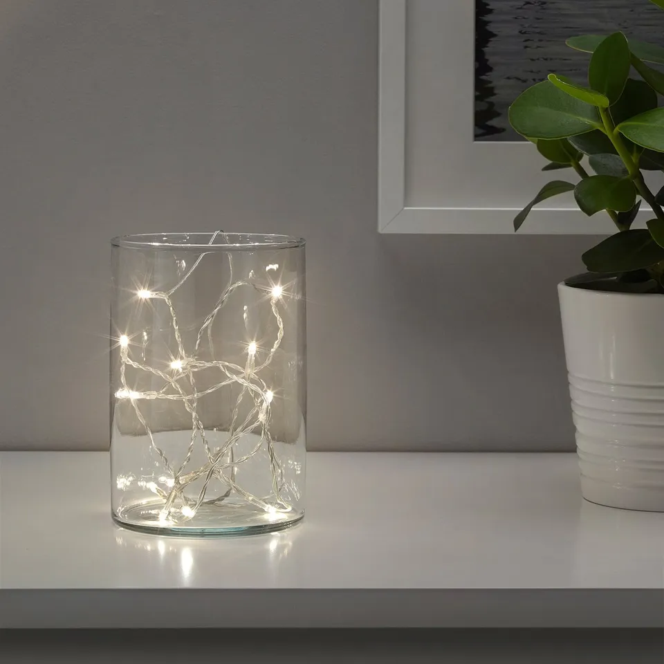 Clear glass vase with led lights inside, emitting warm white light, sits on a white table, accompanied by a white potted plant and a framed photo.