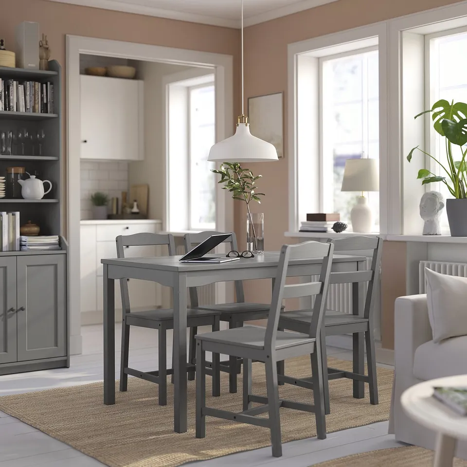 Bright dining area with grey wooden table and chairs, white pendant lamp, jute rug, and bookshelf with accessories, adjacent to a modern kitchen.