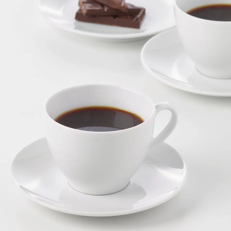 Two white coffee cups on saucers; one with dark coffee, another with chocolate, on a white surface.