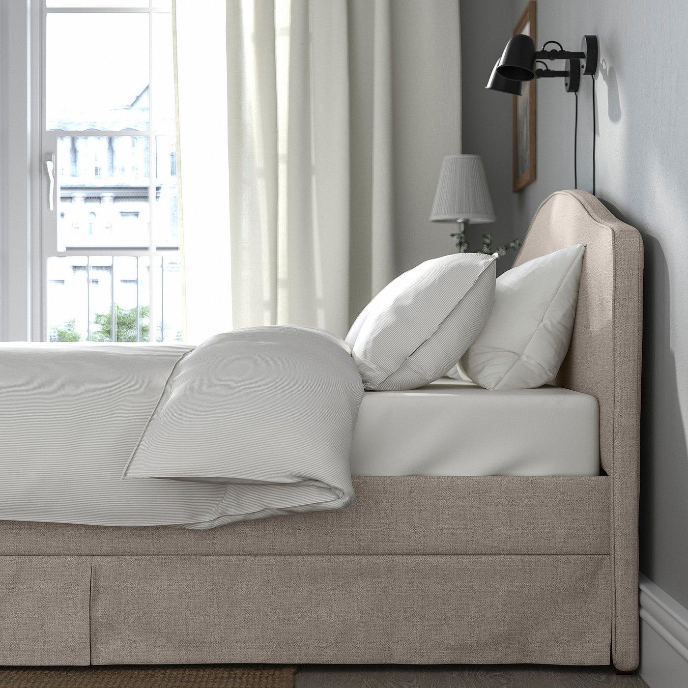 A cosy bed frame with a padded headboard, white pillows, and a folded blanket. A wall-mounted lamp beside it.