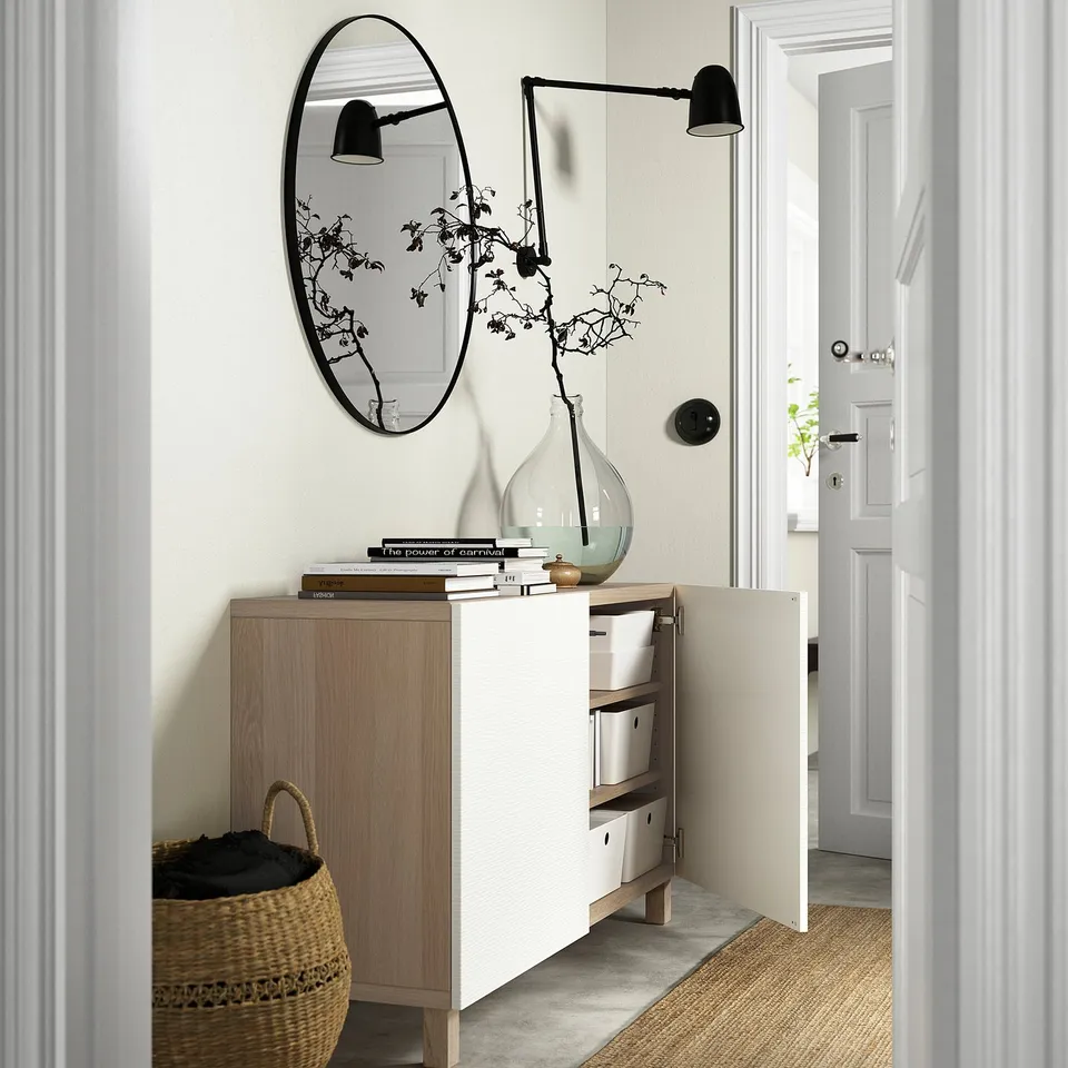 BESTÅ sideboard with adjustable shelves, storing boxes, displaying a vase with branches, books, and illuminated by a wall-mounted light.